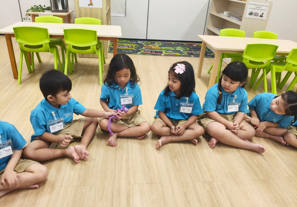 Five children in blue shirts sit on a classroom floor in a circle, passing a purple toy.